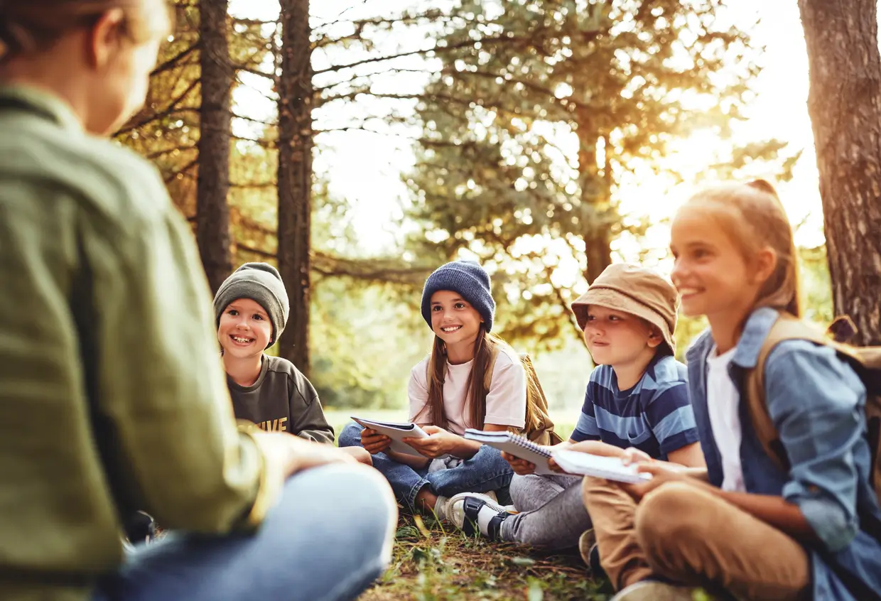 Fünf Kinder sitzen in einem Wald, lächeln und hören einem Erwachsenen zu, während sie Notizbücher halten.