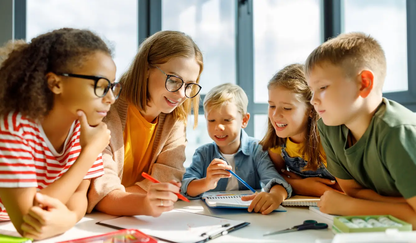 Lehrerin und vier Kinder lächeln und zeichnen gemeinsam an einem Tisch in einem hellen Klassenzimmer. Lehrerin und vier Kinder lächeln und zeichnen gemeinsam an einem Tisch in einem hellen Klassenzimmer.