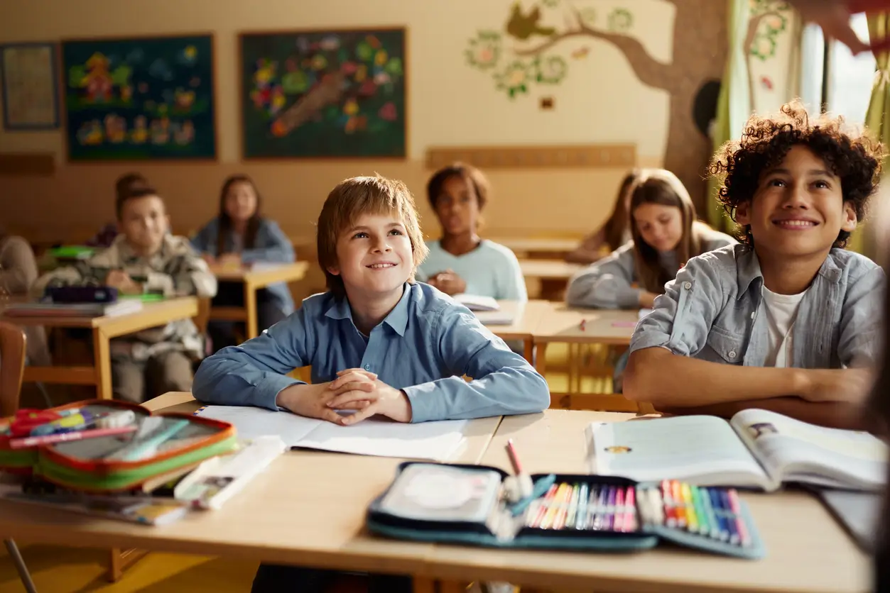 Die Schüler sitzen an ihren Tischen in einem hell erleuchteten Klassenzimmer, lächeln und hören dem Lehrer aufmerksam zu.