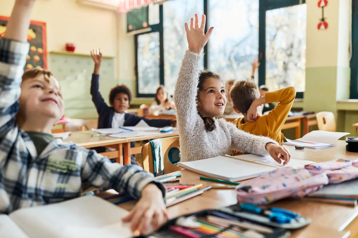 Kinder heben eifrig die Hände in einem Klassenzimmer der Privatschule Schweiz Kosten, Hefte auf den Pulten.
