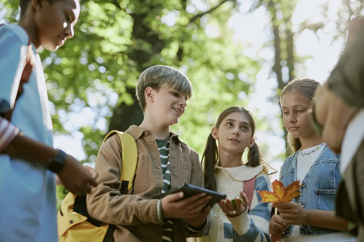 Vier Kinder in einem Park, die sich unterhalten und eine Tafel und Herbstblätter in der Hand halten.