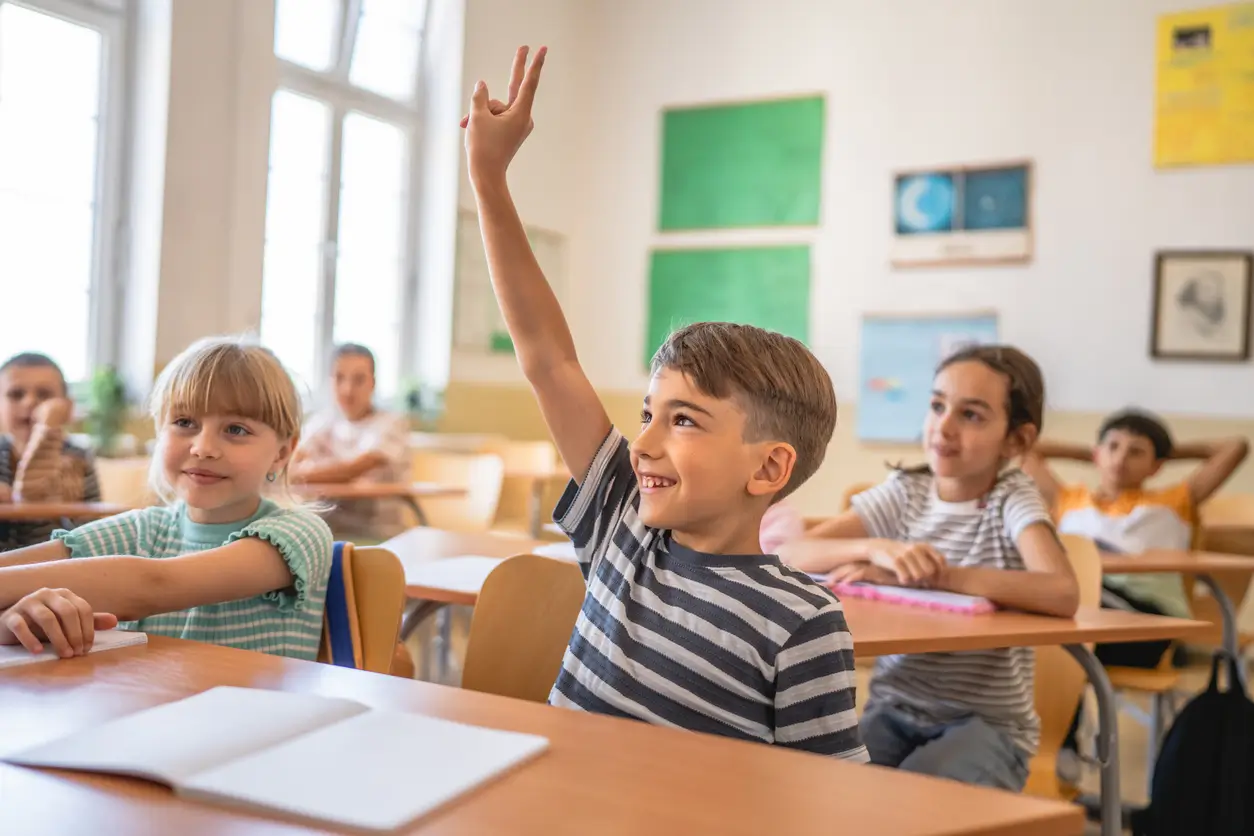 Ein lächelnder Junge hebt seine Hand in einem Klassenzimmer, in dem andere Kinder an Tischen sitzen.