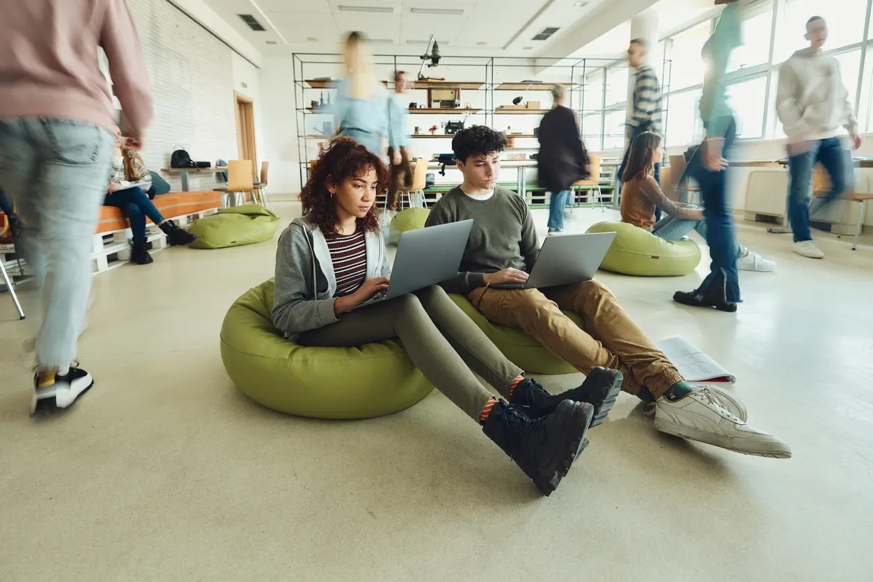 Zwei Schüler sitzen auf Sitzsäcken und arbeiten an Laptops in einem belebten, modernen Klassenzimmer.