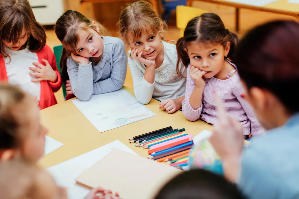 Kleine Kinder sitzen mit Buntstiften an einem Tisch und hören einem Erwachsenen in einem Klassenzimmer zu.