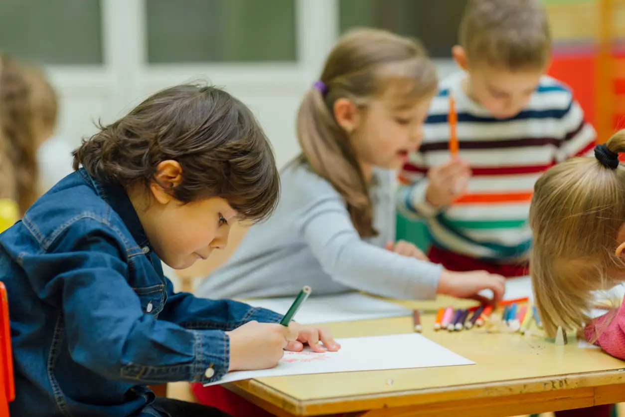 Kinder zeichnen mit Buntstiften an einem Tisch in einem Klassenzimmer.