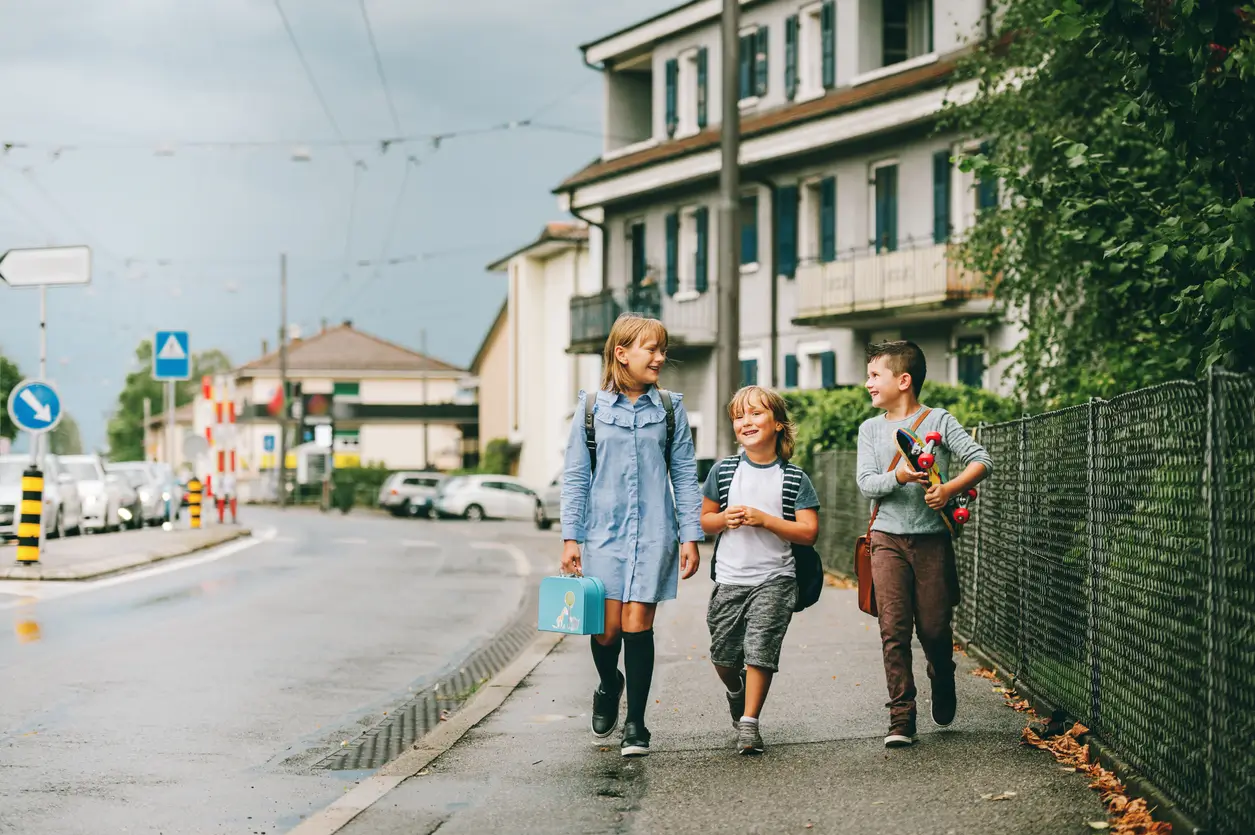 Drei Kinder mit Rucksäcken laufen und unterhalten sich an einem bewölkten Tag auf einem Gehweg in der Nähe von Häusern. Drei Kinder mit Rucksäcken laufen und unterhalten sich an einem bewölkten Tag auf einem Gehweg in der Nähe von Häusern.